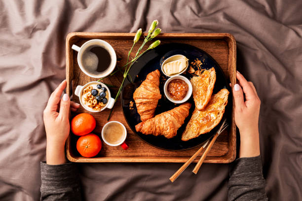 Woman serving continental breakfast on rustic wooden tray in bed (grey sheets). Coffee, croissants, jam, butter, fruits and flowers. Lazy, romantic, weekend morning meal in a hotel suite or home.